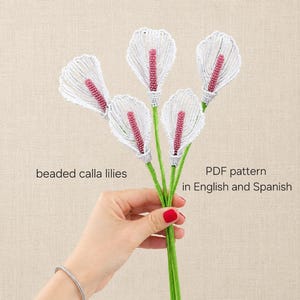 May include: Five beaded calla lilies with white petals and pink centers on green stems. The text reads: beaded calla lilies, PDF pattern in English and Spanish. These decorative floral items are crafted with small beads to form the flower shapes.