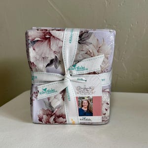May include: A fabric bundle wrapped with a white ribbon featuring the Riley Blake Designs logo. The fabric has a floral pattern in shades of pink, purple, and white. A tag with a photo is attached.