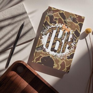 Op de afbeelding: Een rechthoekig notitieboek met harde kaft voorzien van een bruin, beige en donkergroen camouflagepatroon. Het midden toont een wit cirkelvormig spetterontwerp met een groot, gestileerd monogram van de letters TBK, bekroond met een kleine perzikkleurige strik.