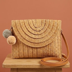 Könnte beinhalten: Gewebte Stroh-Clutch mit Klappenverschluss und einem beigefarbenen Schulterriemen aus Leder. Die Tasche hat ein dekoratives halbkreisförmiges Design auf der Vorderseite und ist mit zwei Pompons in Grau und Beige verziert. Die Tasche steht auf einer Holzoberfläche.