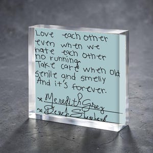 Puede incluir: Un bloque acrílico transparente con una inserción azul claro con texto manuscrito. El texto dice: "Ámense incluso cuando nos odiemos, sin huir... Y es para siempre." Firmado por "Meredith Grey" y "Derek Shepherd."