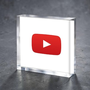 Puede incluir: Un bloque acrílico transparente con el logotipo de YouTube. El logotipo es un rectángulo rojo con esquinas redondeadas y un botón de reproducción blanco. El bloque se muestra sobre una superficie gris oscura.