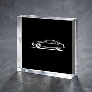 Puede incluir: Un bloque acrílico transparente con un dibujo lineal blanco de un coche clásico sobre un fondo negro. El coche es un cupé con un perfil bajo y un diseño de rueda distintivo. El bloque se muestra sobre una superficie gris.