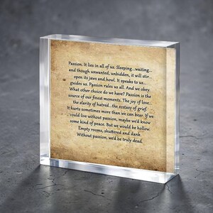 Puede incluir: Un bloque acrílico transparente encierra un papel de estilo vintage con texto impreso. El texto, en una fuente serif clásica, trata sobre la pasión, el amor y el dolor. El bloque rectangular se asienta sobre una superficie oscura, creando una estética moderna.