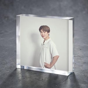 Puede incluir: Un bloque acrílico transparente que muestra una fotografía de una persona con una camiseta blanca. La persona sonríe y tiene la mano en la cadera. El fondo es de color claro.