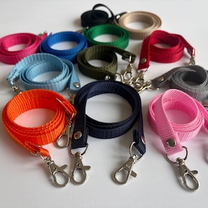 May include: A collection of colorful lanyards in various shades, including pink, blue, orange, and green. Each lanyard is made of a woven material and features a metal clasp and a metal rivet. The lanyards are arranged on a white surface.