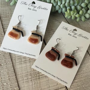 May include: Two pairs of coffee pot earrings on white cards. The earrings are brown, black, and beige, with silver-colored hooks. The cards have the text "The Clay Grotto Est. 2011" and a website address.