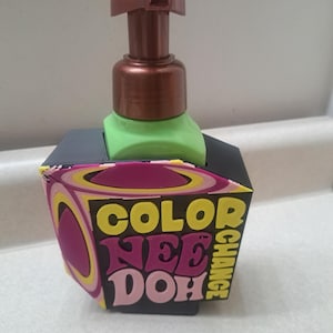 May include: A soap dispenser with a metallic bronze pump and a light green bottle. The bottle is encased in a black silicone sleeve featuring a swirling yellow and magenta pattern and the text COLOR NEE DOH CHANGE in bold, raised lettering.
