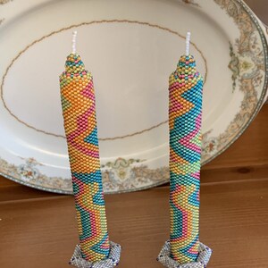 Peut inclure: Deux bougies en perles avec un motif géométrique coloré dans des tons de rose, jaune, bleu et vert. Les bougies sont sur des bougeoirs argentés avec des accents bleus.