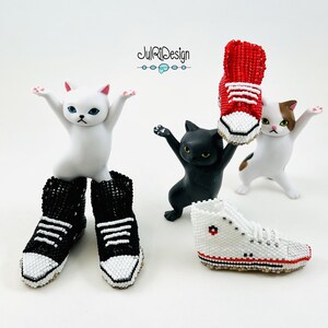 Peut inclure: Trois figurines de chat en c&eacute;ramique blanche avec des baskets en perles rouges, noires et blanches. La basket rouge est tenue en l'air par le chat blanc avec des taches brunes. Le chat noir tient la basket blanche. Le chat blanc aux yeux bleus porte des baskets en perles noires et blanches.