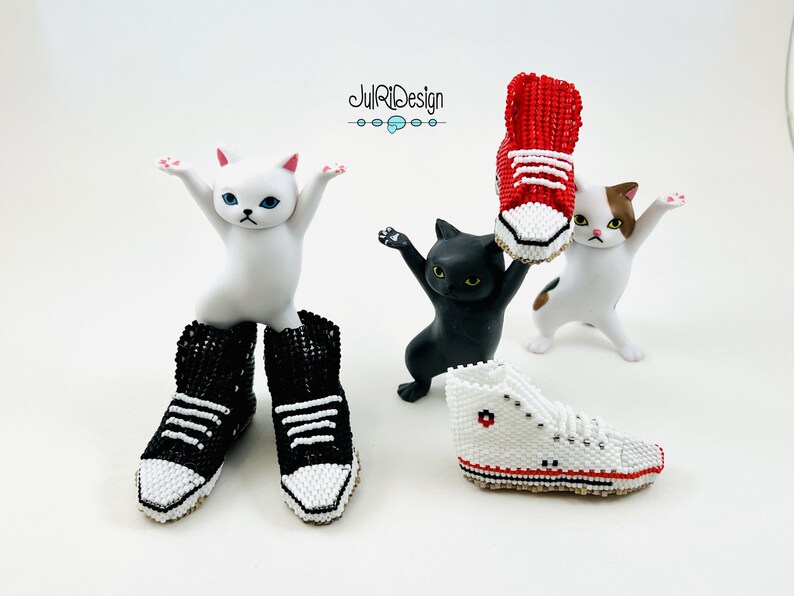 Peut inclure: Trois figurines de chat en c&eacute;ramique blanche avec des baskets en perles rouges, noires et blanches. La basket rouge est tenue en l'air par le chat blanc avec des taches brunes. Le chat noir tient la basket blanche. Le chat blanc aux yeux bleus porte des baskets en perles noires et blanches.