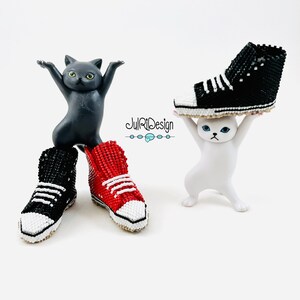 Peut inclure: Deux paires de baskets en perles, une rouge et une noire, sont tenues par deux petites figurines de chats. La basket noire est tenue par une figurine de chat blanc, et la basket rouge est tenue par une figurine de chat gris. Le texte "JulRiDesign" est imprim&eacute; sur la surface blanche sous les figurines.