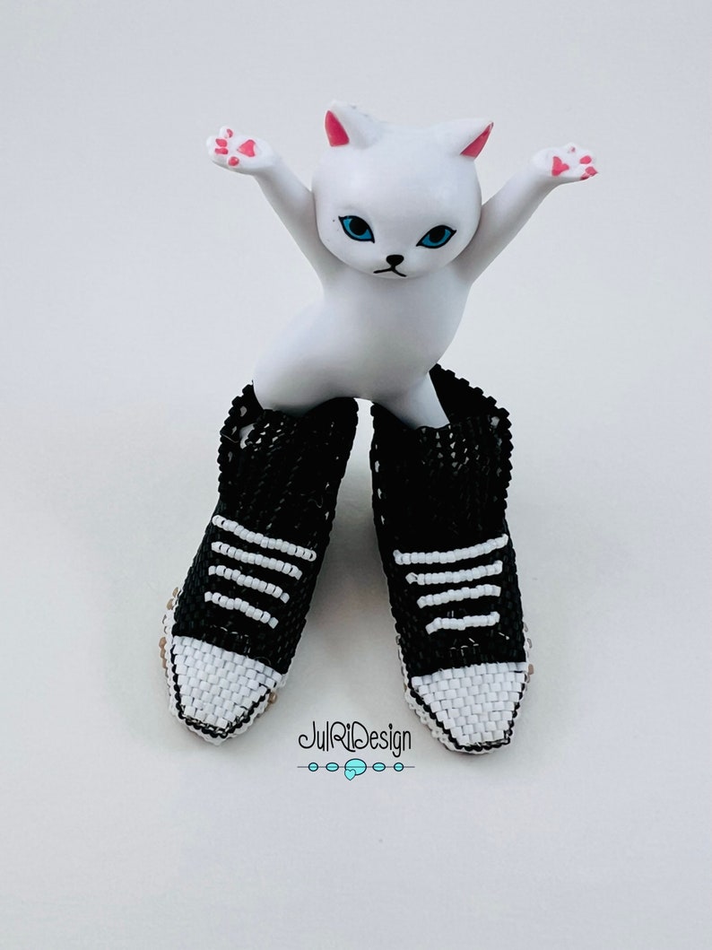 Peut inclure: Une figurine de chat blanc portant une paire de baskets en perles noires et blanches. Les baskets ont des rayures blanches et un bout blanc.