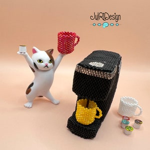 May include: A white cat with brown spots holds a red beaded cup. A black beaded coffee maker with a yellow beaded cup is on a beige surface. There are several small, colorful, beaded coffee pods.