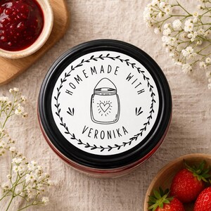 May include: A jar of homemade jam with a black lid and a white label. The label features a jar illustration with a heart and the words "Homemade with Veronika". The jar is surrounded by white flowers and strawberries.