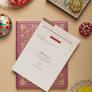 May include: A journal with a purple cover and gold floral design, with a card that reads "YOUR TRANSFORMATION & WHAT'S NEXT". The card also says "A FINAL REMINDER" and "thank you". Decorative boxes and a pomegranate half are also visible.