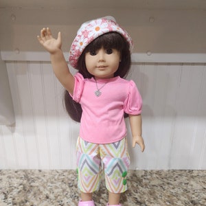 May include: A doll wearing a pink top, patterned capri pants, and pink shoes. The doll has a white hat with a pink floral pattern and is waving. The capri pants have a colorful geometric design.