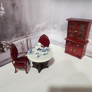 May include: Miniature dollhouse furniture set featuring a round table with a marble top, two red chairs, a red cabinet, and a tea set with blue and white floral designs. The scene is set against a vintage-style backdrop.