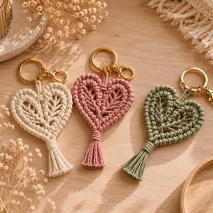Può includere: Tre portachiavi in macramè a forma di cuore nei colori crema, rosa antico e verde salvia. Ogni portachiavi presenta un anello e un moschettone in metallo dorato, intricati motivi a corda annodata e una piccola nappa sulla punta del cuore.