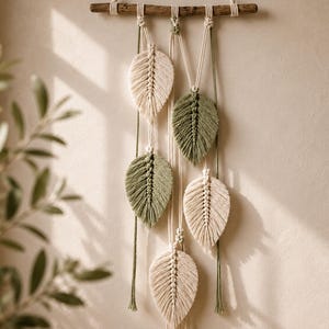 May include: A macrame wall hanging featuring five leaf-shaped fibre elements suspended from a horizontal wooden branch. The leaves are crafted in alternating cream and sage green colours, attached with knotted cords that hang at varying lengths.