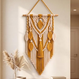 Puede incluir: Un tapiz de macramé con cuerdas de algodón en color amarillo mostaza y crema, suspendido de un soporte de madera. El diseño presenta nudos intrincados, un aro de madera central, varios elementos de flecos en forma de hoja y una borla larga.