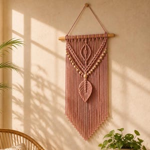 May include: A dusty rose macrame wall hanging featuring intricate knotted patterns, a central leaf-shaped element, and wooden beads suspended from a horizontal wooden dowel with a string loop for mounting on a wall surface.