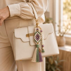 Puede incluir: Un bolso de color crema con un llavero de macram&eacute;. El llavero incluye un nudo de macram&eacute; con patr&oacute;n de diamante en verde salvia y lavanda, un dije de hoja en tono dorado y una hilera vertical de cuentas blancas con el nombre OLIVIA.