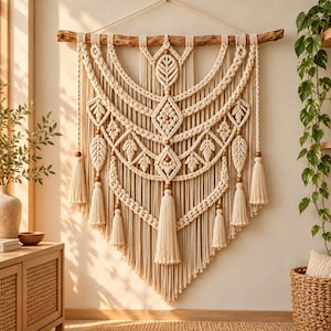 Könnte beinhalten: Ein großer cremefarbener Makramee-Wandbehang, der an einem natürlichen Holzast befestigt ist. Das Design zeigt komplexe Knotenmuster, blattförmige Motive und mehrere hängende Quasten, die mit glatten, runden Holzperlen verziert sind.