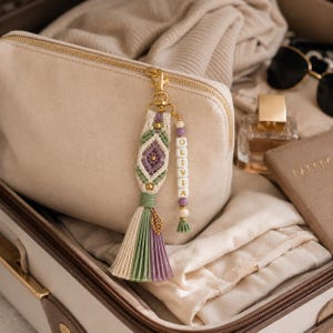 Puede incluir: Un llavero de macram&eacute; con un patr&oacute;n de rombos en cord&oacute;n color crema, verde salvia y lavanda, decorado con cuentas doradas y un dije de hoja. Una hilera vertical de cuentas c&uacute;bicas blancas forma el nombre OLIVIA, con una borla al final.