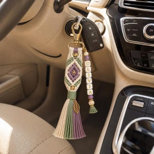 Puede incluir: Un llavero de macram&eacute; con un patr&oacute;n de diamante en cord&oacute;n color crema, verde salvia y morado, decorado con cuentas doradas y un dije de hoja. Una hebra secundaria incluye cuentas de letras blancas que forman OLIVIA con cuentas de madera.
