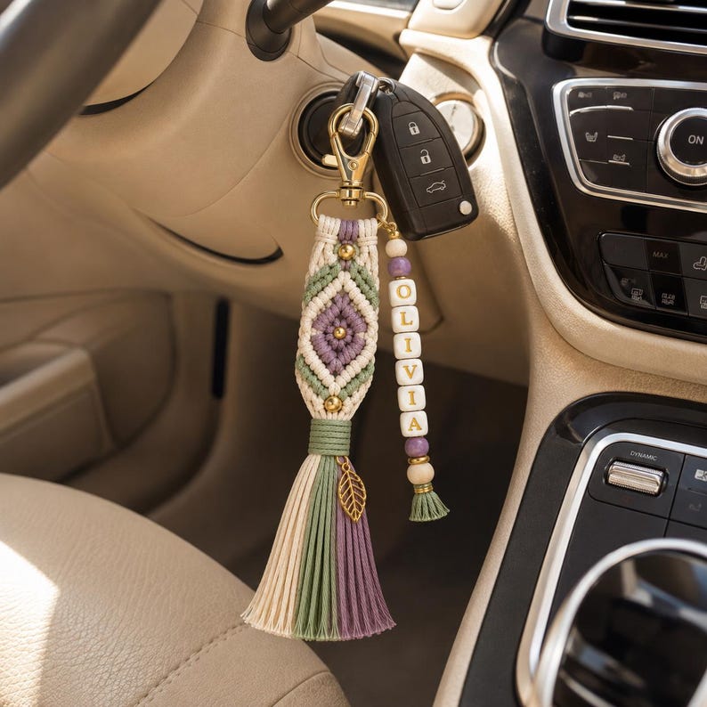 Puede incluir: Un llavero de macram&eacute; con un patr&oacute;n de diamante en cord&oacute;n color crema, verde salvia y morado, decorado con cuentas doradas y un dije de hoja. Una hebra secundaria incluye cuentas de letras blancas que forman OLIVIA con cuentas de madera.