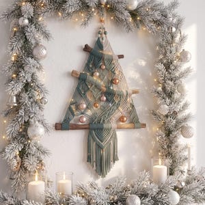 Puede incluir: Un árbol de Navidad de macramé para pared hecho con cuerda de algodón en color verde salvia y crema, con tres ramas de madera horizontales, adornos metálicos pequeños y dijes en forma de hoja, colgado sobre una pared blanca con follaje.