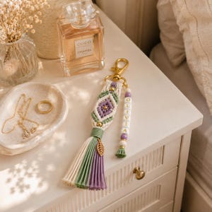 Puede incluir: Un llavero de macram&eacute; con cord&oacute;n de algod&oacute;n en color crema, verde salvia y lavanda en un patr&oacute;n de rombos con un cierre de metal dorado. Una tira colgante incluye cuentas blancas con las letras OLIVIA, cuentas lavanda y un dije de hoja dorada.