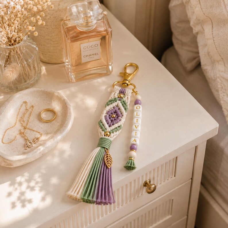 Puede incluir: Un llavero de macram&eacute; con cord&oacute;n de algod&oacute;n en color crema, verde salvia y lavanda en un patr&oacute;n de rombos con un cierre de metal dorado. Una tira colgante incluye cuentas blancas con las letras OLIVIA, cuentas lavanda y un dije de hoja dorada.