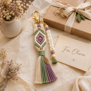 Puede incluir: Un llavero de macram&eacute; con patr&oacute;n de diamante en cord&oacute;n color crema, verde salvia y lavanda con cuentas doradas. Un hilo lateral incluye cuentas de letras blancas que forman OLIVIA y un dije de hoja dorada. La borla es crema, verde y morada.