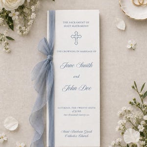 May include: A white wedding programme featuring blue text and a cross icon, tied with a dusty blue chiffon ribbon. The text reads: THE SACRAMENT OF HOLY MATRIMONY, THE CROWNING IN MARRIAGE OF Jane Smith and John Doe, SATURDAY, THE TWENTY-SIXTH of JUNE two thousand twenty-six, Saint Barbara Greek Orthodox Church.