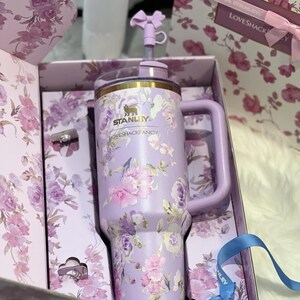 May include: A lavender Stanley tumbler with a floral design, a handle, and a straw. The tumbler is in a gift box with a matching floral pattern. The Stanley logo and the words "LOVESHACKFANCY" are visible on the tumbler.