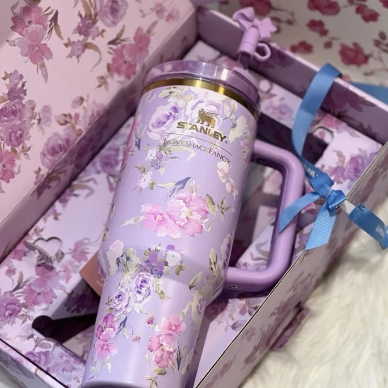 May include: A lavender Stanley tumbler with a floral design, featuring pink and purple flowers. The tumbler has a handle and a lid, and is presented in a matching floral-patterned box with a blue ribbon.