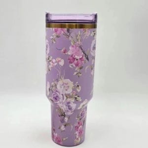May include: A lavender tumbler with a floral design featuring pink and white flowers. The tumbler has a clear lid and a gold-colored band near the top. The overall design is elegant and feminine.