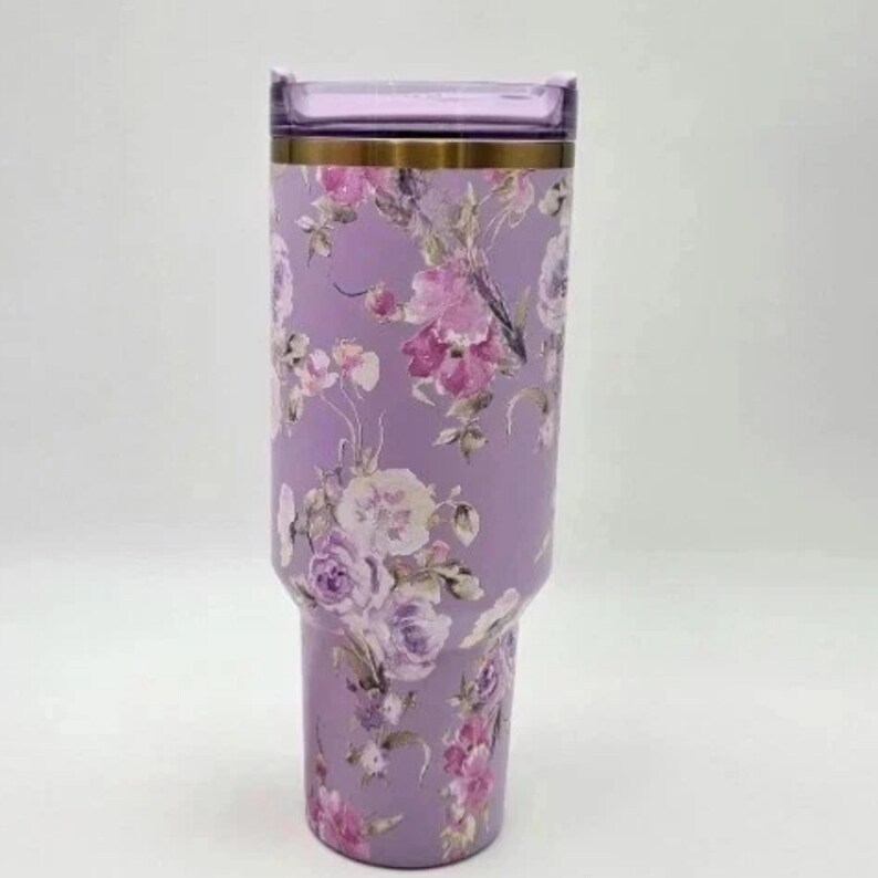 May include: A lavender tumbler with a floral design featuring pink and white flowers. The tumbler has a clear lid and a gold-colored band near the top. The overall design is elegant and feminine.