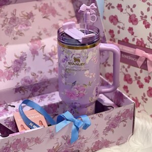 May include: A lavender Stanley mug with a floral design, a handle, and a lid. The mug is inside a box with a matching floral pattern and a blue ribbon. The text "LOVESHACKFANCY" is visible on the mug.