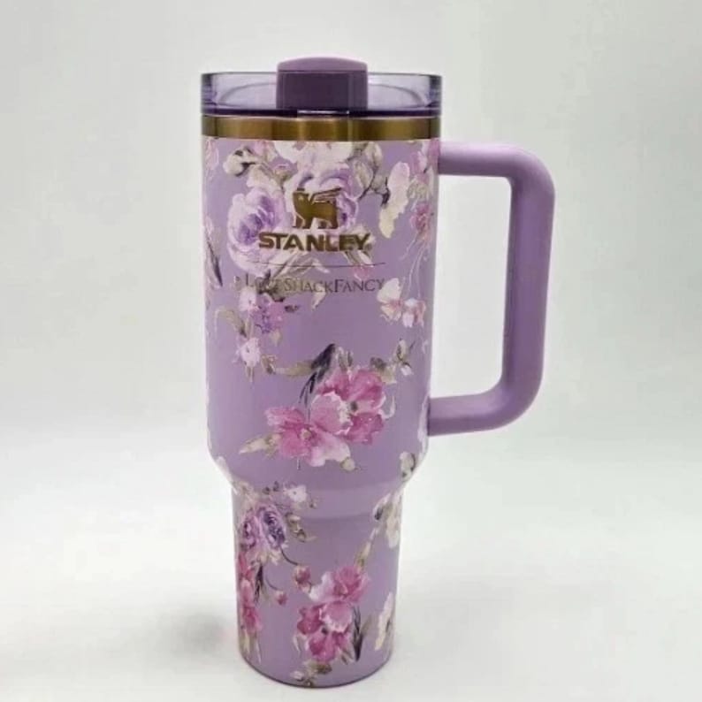 May include: A purple Stanley tumbler with a floral design. The tumbler has a handle, a lid, and the text "STANLEY" and "SHACKFANCY". The floral pattern includes pink and purple flowers.