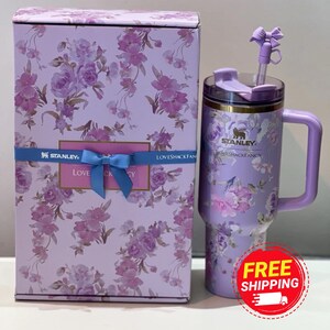 May include: A floral-patterned Stanley tumbler and matching gift box. The tumbler is lavender with a handle, lid, and straw. The box is lavender with a floral design and a blue ribbon. The tumbler has the text "LOVESHACKFANCY".
