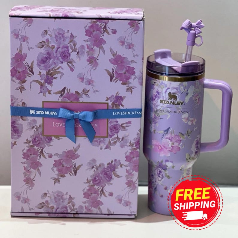 May include: A floral-patterned Stanley tumbler and matching gift box. The tumbler is lavender with a handle, lid, and straw. The box is lavender with a floral design and a blue ribbon. The tumbler has the text "LOVESHACKFANCY".