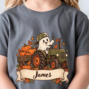 Puede incluir: Una camiseta gris carbón con un gráfico de un fantasma blanco con gorra de béisbol verde, conduciendo un tractor verde que tira de un remolque lleno de calabazas naranjas y hojas de otoño, con una pancarta debajo que muestra el nombre James.