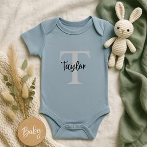 Könnte beinhalten: Hellblauer Baby-Body mit dem Namen "Taylor" in schwarzer Schrift unter einem großen grauen Buchstaben "T". Ein kleines, cremefarbenes Häschen aus Häkelarbeit und eine Holzscheibe mit dem Wort "Baby" sind ebenfalls im Bild.