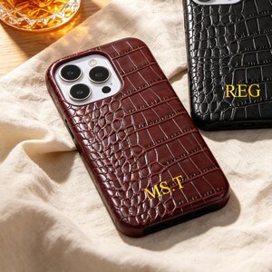 Puede incluir: Tres fundas para smartphone con un patrón de textura de piel de cocodrilo. Una funda es de color rojo oscuro con letras doradas MS.T, una es negra con letras doradas REG y una es crema con letras doradas RS. Cada funda tiene un recorte triple cámara.