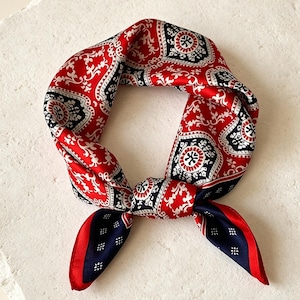 Puede incluir: Un pañuelo de seda rojo, blanco y azul marino con un estampado de cachemira. El pañuelo está anudado y atado, con los extremos colgando. El diseño del pañuelo presenta un patrón repetitivo de formas florales y geométricas.