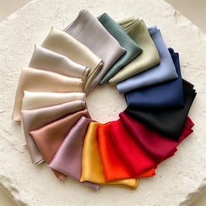 May include: A collection of folded silk pocket squares in a circular arrangement. The pocket squares display a spectrum of colors, including beige, green, blue, black, red, and yellow. The fabric has a subtle sheen, suggesting a luxurious texture.