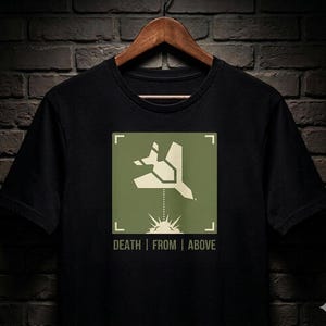 May include: Black t-shirt featuring a green square graphic with a stylized bird design and the text "DEATH | FROM | ABOVE". The design includes a dotted line and a sunburst-like element. The shirt is hanging on a wooden hanger.